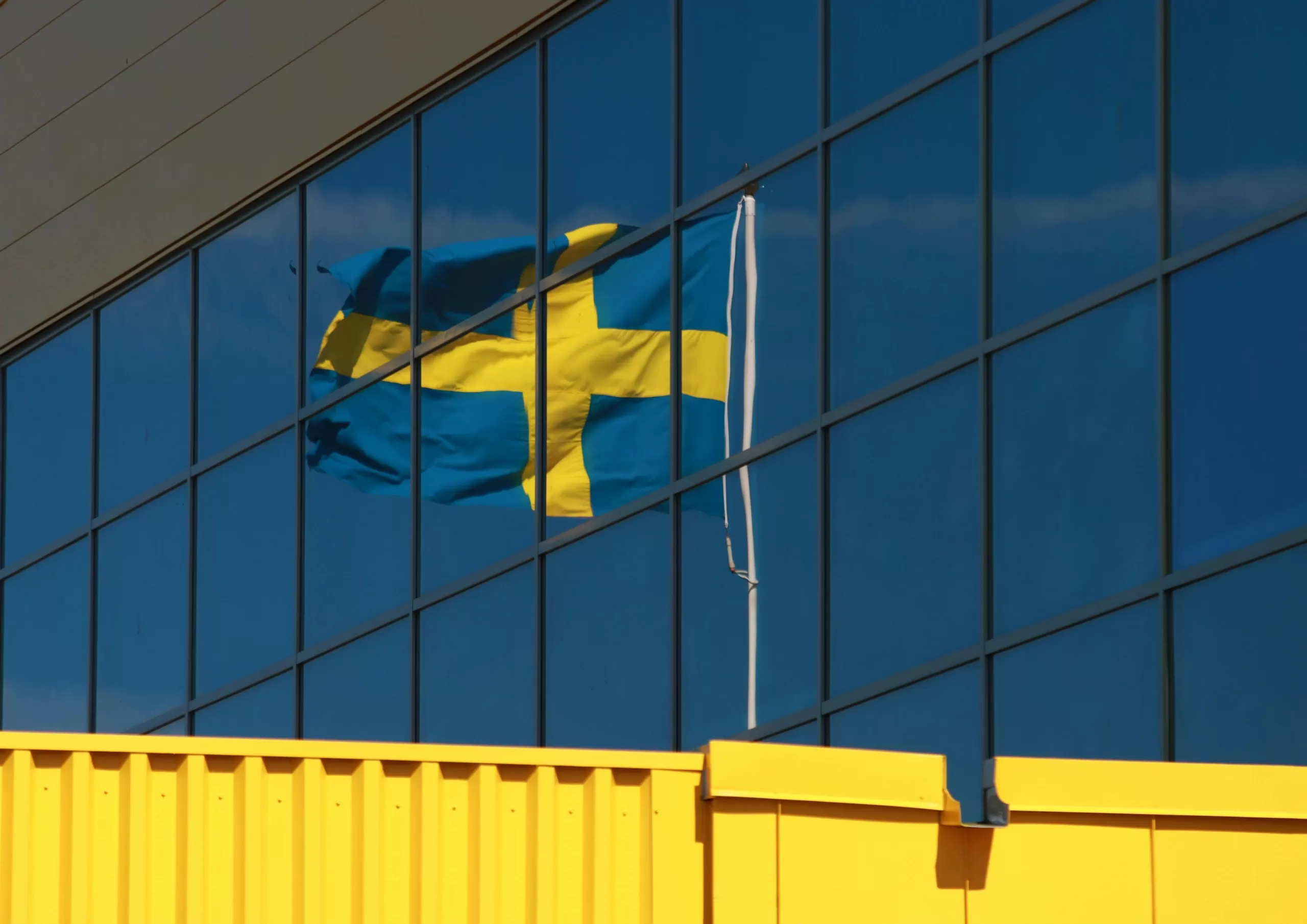 Bendera Swedia berkibar di depan gedung kaca, visual pendukung tema brand Swedia dan kualitas desain.