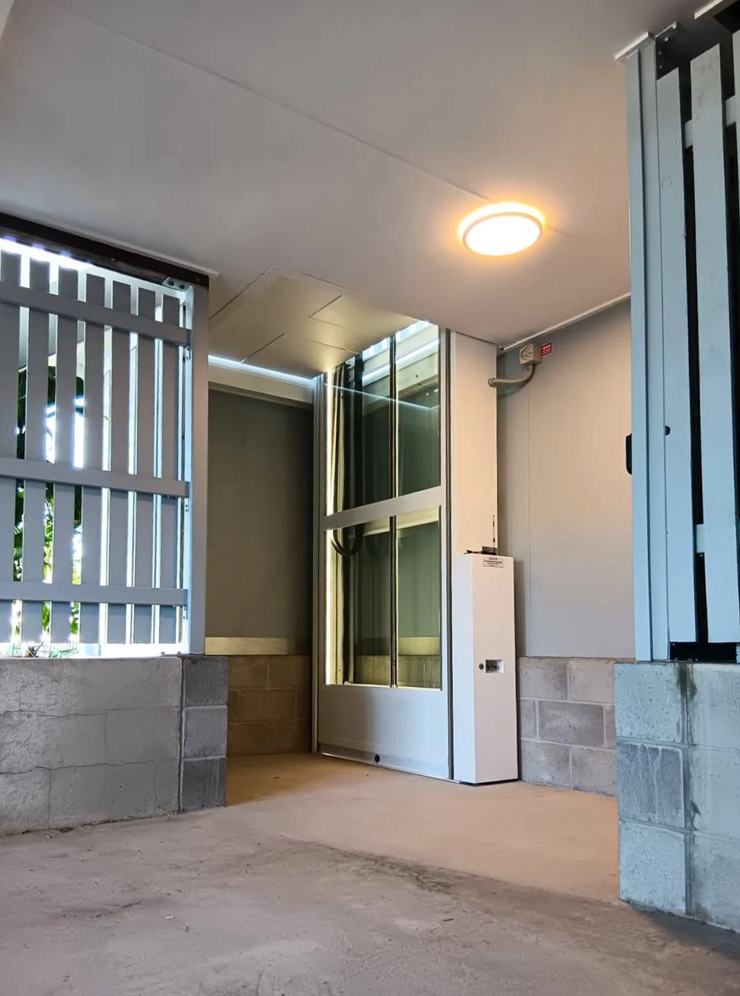Mencari Outdoor Lift? Kenali Solusi Lift Rumah Semi-Outdoor yang Tepat 11 Lift rumah platform terpasang di area semi-outdoor beratap dengan dinding pelindung dan struktur bangunan di sekelilingnya.