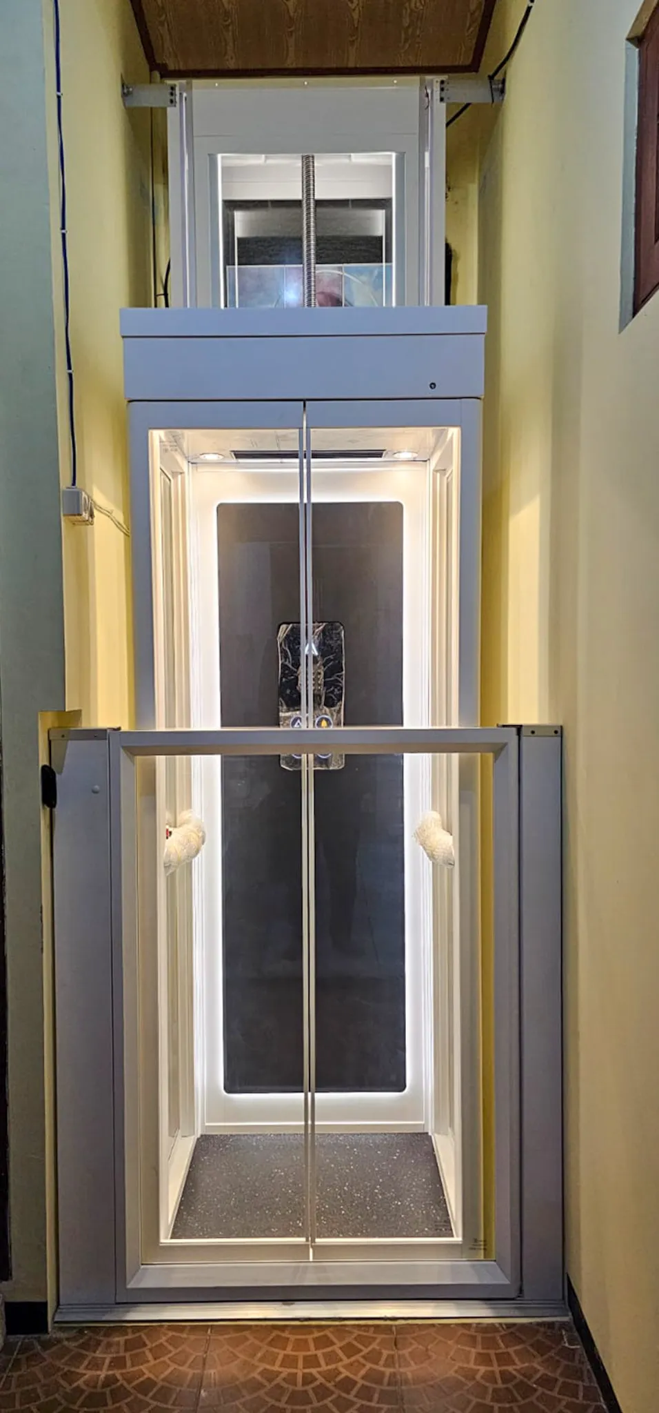 Lift Rumah Terkecil di Indonesia | Ascenda Size S 12 Lift rumah kecil Ascenda Size S dengan pintu kaca dan kabin berwarna putih di lorong sempit.